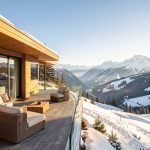 Chalet contemporain en montagne avec vue alpine, terrasse ensoleillée et ambiance hiver lumineuse, tons chauds de bois et pierre dans un paysage neigeux haut de gamme.