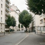 Vue urbaine réaliste de Villeurbanne montrant des immeubles d’habitation, des arbres, des trottoirs calmes et quelques panneaux d’annonces vierges dans une ambiance documentaire sobre.